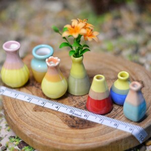 May include: A collection of miniature ceramic vases in various colors, including yellow, pink, blue, and red, arranged on a wooden slice. Some vases hold small orange flowers. A measuring tape is placed across the wooden slice.