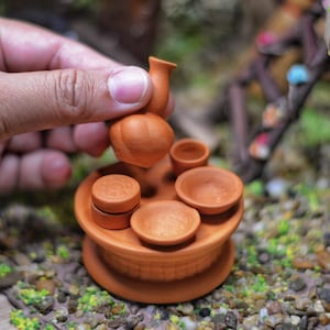 Puede incluir: Un conjunto de terracota en miniatura con una jarrita pequeña y cuatro tazas, dispuestas sobre una base redonda. El conjunto está hecho de arcilla marrón rojiza y tiene un aspecto rústico y hecho a mano.