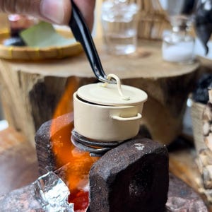 Pode incluir: Uma panela branca em miniatura com uma tampa está sendo segurada sobre um pequeno fogão improvisado com uma chama. O fogão é feito de um material marrom escuro e tem um pedaço de papel alumínio no lado.