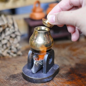 May include: A small, brass-colored pot with a lid, sitting on a black metal stand with a small fire burning inside. The pot is being opened by a hand. A stack of firewood is in the background.