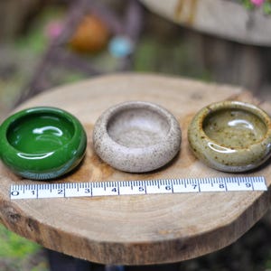 May include: Three small, round ceramic bowls in green, beige, and tan are displayed on a wooden slice. A ruler is placed near the bowls for scale. The bowls have a smooth, glossy finish and are likely for decorative or miniature purposes.