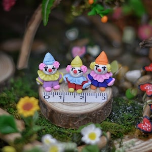 May include: Three miniature clown figurines with colorful outfits and hats, standing on a wooden slice with a ruler. The clowns have red noses and painted faces. The scene is set in a miniature garden with moss and flowers.
