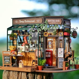 Puede incluir: Modelo en miniatura de madera de un bistró-bar y restaurante con un toldo verde. Los letreros dicen "Tipsy Restaurant" y "Bistro Bar". El modelo incluye un oso de peluche, mesas, sillas y plantas en miniatura. Un letrero anuncia "Cerveza".