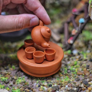 May include: A miniature terracotta teapot with a lid pouring tea into four small terracotta cups on a round terracotta tray.