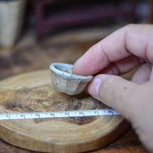 May include: A small, handmade ceramic bowl with a light beige speckled finish and a white rim. The bowl is held over a wooden cutting board with a ruler, suggesting a measurement of the bowl's size.