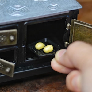 Pode incluir: Um forno preto em miniatura com uma placa de fogão prateada e duas portas abertas. No interior, uma pequena grelha preta contém duas batatas amarelas. Uma mão está a entrar para ajustar a grelha.