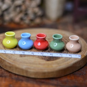 Może przedstawiać: Pięć miniaturowych ceramicznych wazonów w kolorze żółtym, niebieskim, czerwonym, zielonym i brzoskwiniowym, ułożonych na drewnianym plastrze. Linijka umieszczona przed wazonami. Wazony mają zaokrąglony kształt z małym otworem.