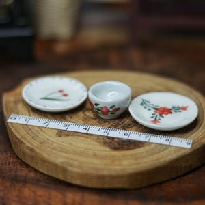 Puede incluir: Juego de té de cerámica en miniatura con diseños florales, que incluye dos platos pequeños y una taza, dispuestos sobre un posavasos de madera. Una regla se coloca sobre el posavasos, mostrando las medidas.