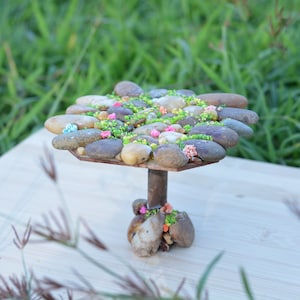 Puede incluir: Una mesa pequeña hecha de piedras con una base marrón y una parte superior decorada con piedras pequeñas, musgo verde y flores rosas y azules.