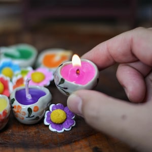 Könnte beinhalten: Eine brennende, herzförmige rosa Kerze, die von einer Hand gehalten wird, umgeben von anderen bunten Kerzen in blumen- und herzförmigen Keramikbehältern. Die Kerzen sind in verschiedenen Farben, darunter Blau, Gelb und Lila.