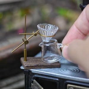 Może przedstawiać: Miniaturowy szklany zestaw do parzenia kawy z przezroczystą szklaną karafką i żebrowanym stożkiem filtrującym. Zestaw jest podparty mosiężnym stojakiem na drewnianej podstawie, umieszczonym na miniaturowej kuchence. Do domków dla lalek.