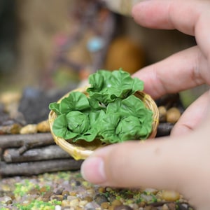 Pode incluir: Uma cesta de vime em miniatura cheia de alface verde. A cesta é segurada por uma mão, destacando a pequena escala do alimento. A cena sugere uma casa de bonecas ou uma exposição de comida em miniatura.