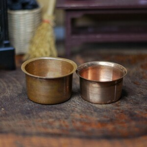 Puede incluir: Dos pequeños recipientes redondos de metal color cobre sobre una superficie de madera. Los recipientes tienen una forma cilíndrica simple con un borde ligeramente ensanchado. La iluminación resalta el brillo metálico del cobre.