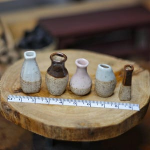 Może przedstawiać: Kolekcja pięciu miniaturowych ceramicznych wazonów w różnych kształtach i kolorach, w tym białym, brązowym i różowym. Wazony ułożone są na drewnianym plastrze z linijką dla skali. Wazony mają około 2,5 do 5 cm wysokości.