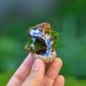Puede incluir: Escena de cascada de jardín de hadas en miniatura. El pequeño y detallado diorama presenta una cascada con agua azul cayendo en cascada sobre rocas cubiertas de musgo verde. Pequeñas flores rosas añaden color a la escena. La base es una pequeña maceta redonda y moteada.