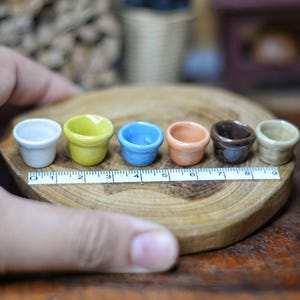 May include: A collection of six miniature ceramic pots in various colors, including white, yellow, blue, orange, brown, and beige. The pots are arranged on a wooden slice with a ruler for scale. These small pots are ideal for miniature gardening or crafts.