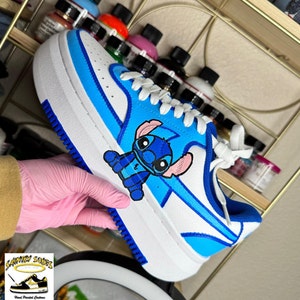 May include: White sneakers with blue accents and a hand-painted cartoon Stitch character. The sneakers have a blue and white stripe design and a blue sole.