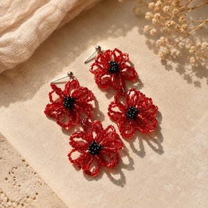May include: A pair of drop earrings featuring three flower-shaped motifs crafted from translucent red seed beads. Each flower has a cluster of small, opaque black beads at its center. The earrings are attached to silver-toned metal posts.