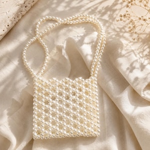 May include: A small, square-shaped handbag constructed entirely from interconnected white faux pearls. The bag features a structured, open-weave lattice pattern and a single integrated handle made from the same pearl bead design.