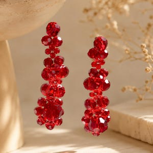 May include: A pair of long, dangling earrings composed of multiple faceted, deep red crystal beads arranged in a cascading, cluster-like formation. The beads vary in size, creating a textured, reflective surface that catches light throughout.