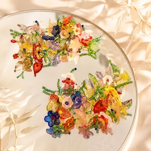 May include: Two hair clips featuring a dense arrangement of multi-colored glass and acrylic flowers in shades of red, blue, yellow, and pink, accented with green beaded stems and leaves. The floral clusters are intricately wired together.
