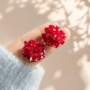 May include: A pair of round stud earrings featuring a cluster of deep red faceted crystals and small matching red beads. The spherical design creates a textured, multi-dimensional appearance with light reflecting off the sharp crystal edges.