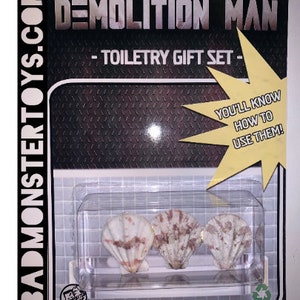 May include: A toy packaging featuring a yellow and black design with the text "Demolition Man - Toiletry Gift Set -" and a picture of three white seashells with brown markings in a clear plastic container. The packaging also includes the text "You'll know how to use them!" and the website address "badmonstertoys.com".