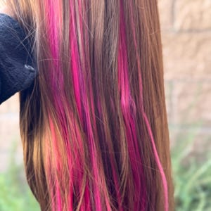 Op de afbeelding: Close-up van lang bruin haar met levendige roze highlights. De roze strepen zijn verspreid over het bruine haar, waardoor een gedurfd en kleurrijk contrast ontstaat. Het haar lijkt steil gestyled te zijn.