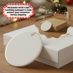 May include: A smooth, circular white ceramic ornament featuring a small hole at the top for hanging. The ornament is presented alongside a plain white gift box, with a secondary view showing a similar ornament resting on a brown cardboard box.