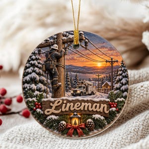 May include: A round hanging ornament featuring a winter scene with a utility worker climbing a wooden pole, a bucket truck, and a snowy landscape at sunset. The word Lineman is written in raised script on a wooden plaque with pine and red berries.