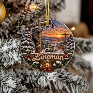 May include: A round Christmas ornament featuring a scene of a utility worker on a wooden pole, a utility truck, and snow-covered pine trees at sunset. The word Lineman is written in script on a wooden sign, accented by pine cones and a red bow.