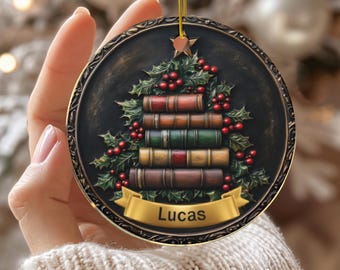 May include: A round, ceramic Christmas ornament with a dark background and a gold-colored trim. The ornament features a stack of books arranged in the shape of a Christmas tree, with holly and red berries. A banner at the bottom reads "Lucas".