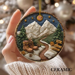 May include: A round ceramic ornament featuring a mountain landscape with hiking boots, trees, and a path. The design includes a night sky with a moon and stars. The word "CERAMIC" is at the bottom.