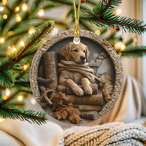 May include: A round Christmas ornament featuring a golden retriever puppy wearing a scarf and sitting on books. The ornament has a detailed relief design with a gold hanging cord and is surrounded by a decorative border. The background includes fall leaves.