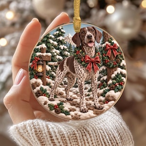 Könnte beinhalten: Ein runder Weihnachtsanhänger mit einem braun-weiß gefleckten Hund, der einen festlichen Kranz mit roter Schleife trägt. Der Hintergrund zeigt eine verschneite Waldszene mit Tannen, einer leuchtenden Laterne an einem Pfosten und einem Kranz.