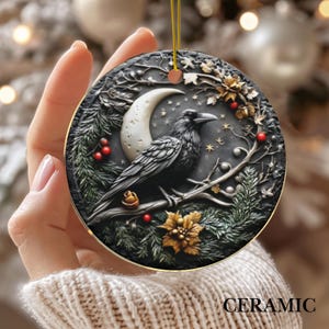 May include: A round ceramic ornament featuring a detailed raven perched on a branch with a crescent moon and stars. The design includes gold accents, red berries, and the word "CERAMIC" at the bottom. The ornament has a gold string for hanging.