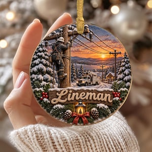 May include: A round ceramic Christmas ornament featuring a winter scene of a utility worker climbing a wooden power pole. The image includes a utility truck, snow-covered pine trees, and a sunset. The word Lineman is printed on a wooden sign.