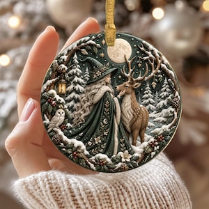 Puede incluir: Un adorno redondo de cerámica con una ilustración detallada de una figura con capa verde oscuro y sombrero puntiagudo en un bosque nevado junto a un ciervo y un búho. La escena incluye una linterna brillante, pinos y una luna llena.