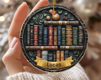 May include: A round ceramic ornament featuring a detailed bookshelf design with various colorful books. The ornament has a gold ribbon for hanging and a banner with the name "Lucas". The word "CERAMIC" is printed at the bottom.