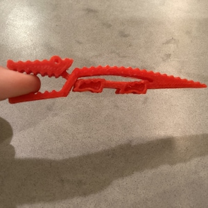 May include: A red plastic alligator-shaped clip. The clip is designed to hold papers or other items together.