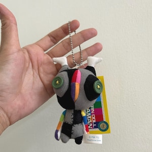 May include: A small, handmade doll with a unique design. The doll has a gray body with black patches, button eyes, and a colorful striped head. It is attached to a silver chain and has a yellow tag with Japanese text.