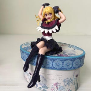 Może przedstawiać: Figurka młodej kobiety z blond włosami, ubranej w ciemnoczerwony i czarny strój oraz czarne buty. Siedzi na ozdobnym pudełku z kwiatowym i niebieskim wzorem.