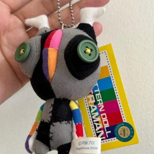 May include: A gray and black plush doll with colorful patchwork details, green button eyes, and white feet. The doll has a silver chain and a yellow tag with Japanese text and a colorful design. The tag also includes the text "PATTERN DOLL ULTRAMAN".