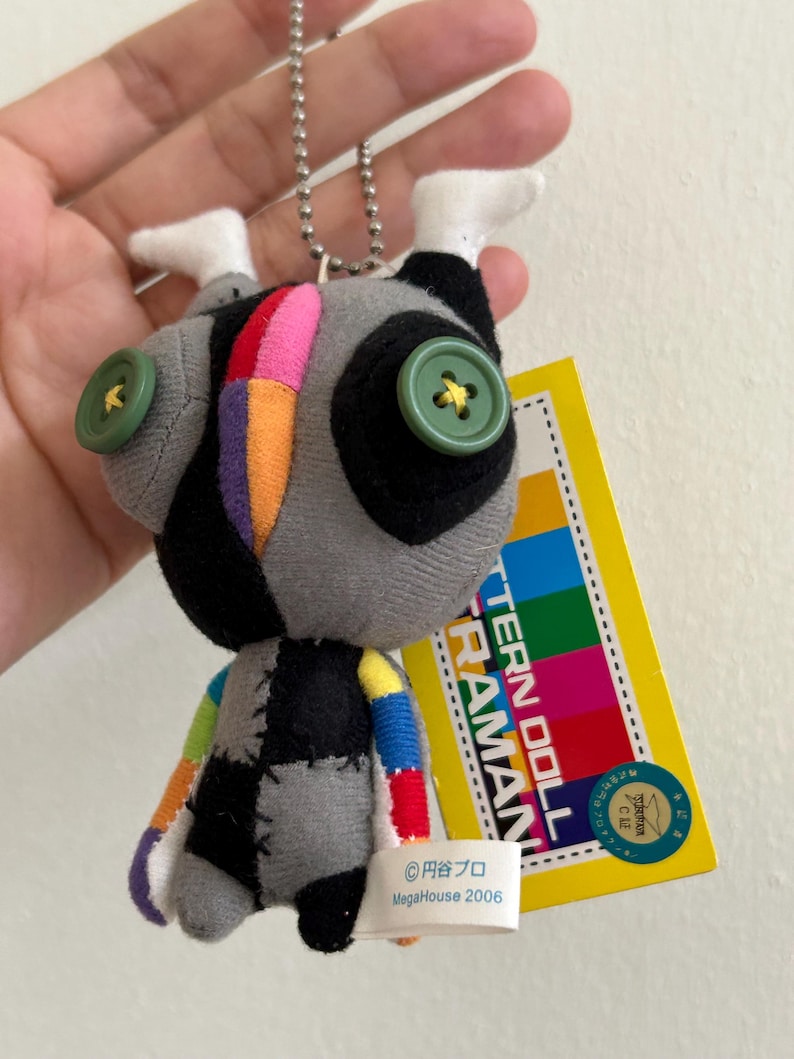 May include: A gray and black plush doll with colorful patchwork details, green button eyes, and white feet. The doll has a silver chain and a yellow tag with Japanese text and a colorful design. The tag also includes the text "PATTERN DOLL ULTRAMAN".