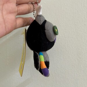 May include: A small, plush keychain in the shape of a stylized bird. The bird is primarily black with gray accents, a green button eye, and a colorful tail. It hangs from a silver chain and has a yellow tag attached.