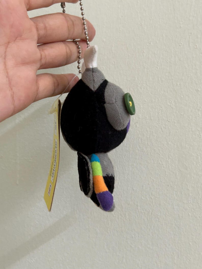 May include: A small, plush keychain in the shape of a stylized bird. The bird is primarily black with gray accents, a green button eye, and a colorful tail. It hangs from a silver chain and has a yellow tag attached.