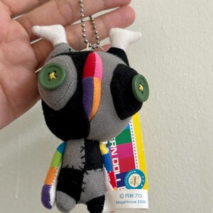 May include: A small, handmade doll with a patchwork design. The doll features a gray body with black and white accents, button eyes, and colorful stitched details. It has a silver chain for hanging and a tag with Japanese text.