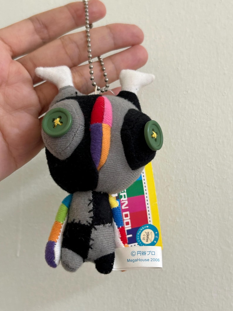 May include: A small, handmade doll with a patchwork design. The doll features a gray body with black and white accents, button eyes, and colorful stitched details. It has a silver chain for hanging and a tag with Japanese text.