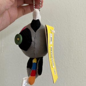 May include: A plush toy with a button eye, a colorful tail, and a metal chain. The toy is primarily gray with black and pink accents. A yellow tag is attached to the toy.