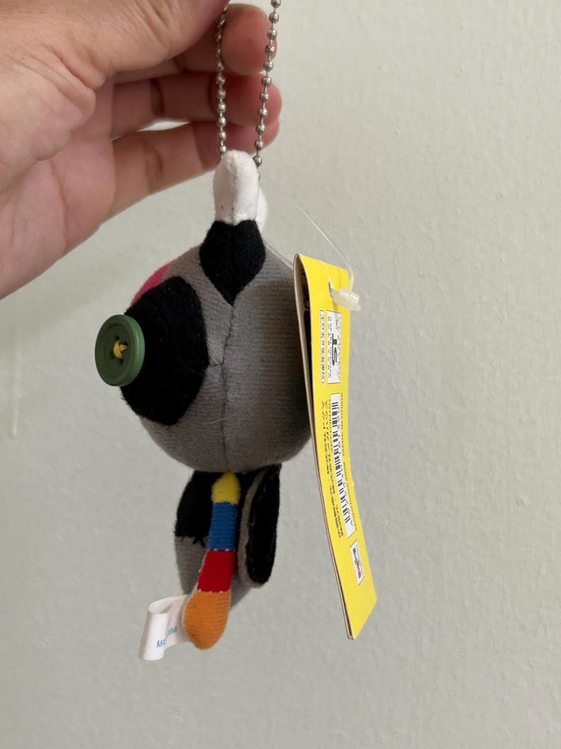 May include: A plush toy with a button eye, a colorful tail, and a metal chain. The toy is primarily gray with black and pink accents. A yellow tag is attached to the toy.